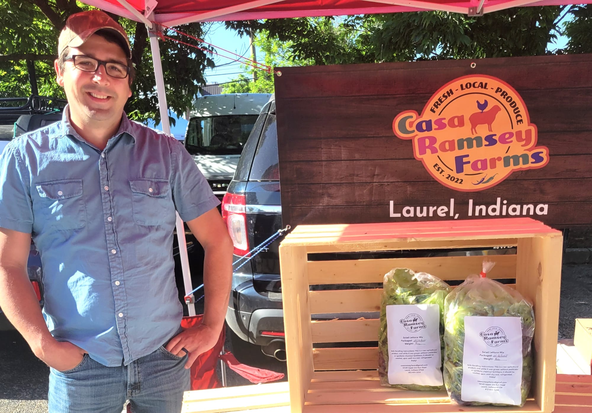 Mark Ramsey stands next to his display at the Oxford Farmers Market, with a sign for Casa Ramsey Farms and two bags of lettuce in a crate