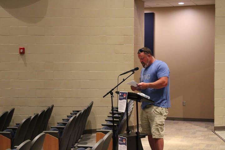 Scotty King reads from a sheet of notes at a microphone in an auditorium