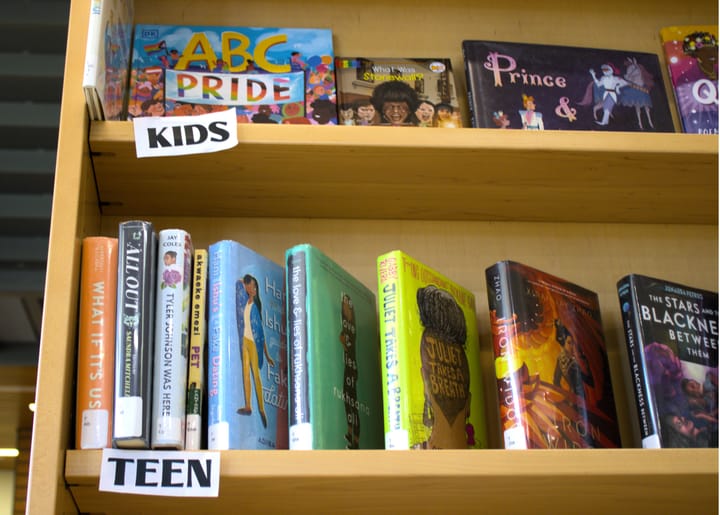 An array of multicolored LGBTQ books on a book shelf, top shelf is labeled kids and the one below it is teens.
